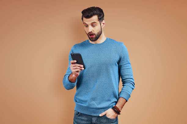 istock Surprised young man in casual wear using digital tablet and making a face while standing against brown background 1224573710