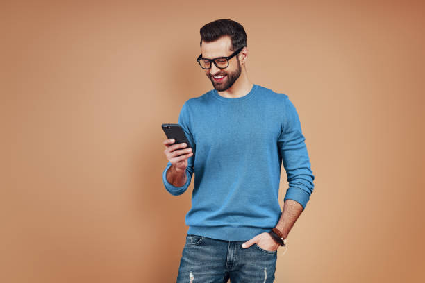 istock Happy young man in smart casual wear using smart phone and smiling while standing against brown background 1224576174