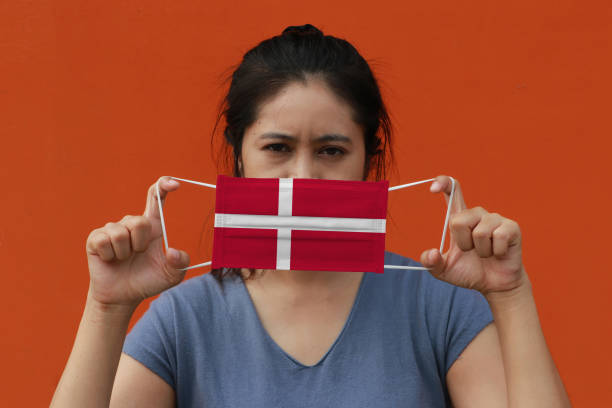 une femme avec le drapeau du danemark sur le masque hygiénique dans sa main et a soulevé le visage avant sur le fond de couleur orange. tiny particle ou virus corona ou covid 19 protection. - arms lifted photos et images de collection