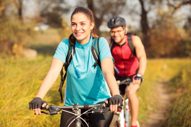 Happy young woman riding mountain bike with her boyfriend Beautiful happy young Caucasian woman riding mountain bike over a meadow in the early morning and her boyfriend is following. Healthy and active sporty lifestyle red white blue bike stock pictures, royalty-free photos & images