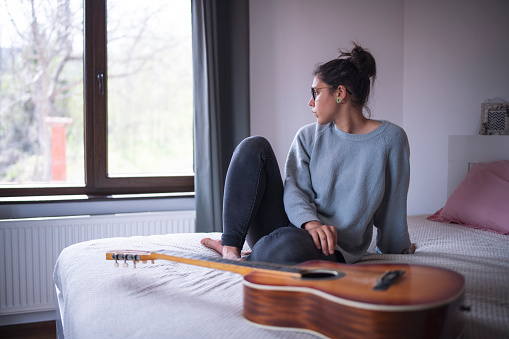 Depressed woman sitting at home and looking through the window, he standing in front of acoustic guitar.