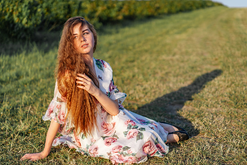 Full length portrait of beautiful woman with long curly hair on currant field background. Girl in a light dress sits on the grass in sunny day.