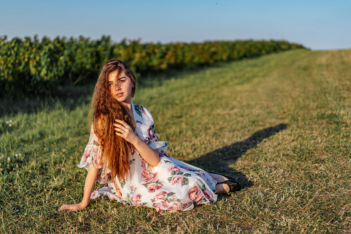 Full length portrait of beautiful woman with long curly hair on currant field background. Girl in a light dress sits on the grass in sunny day.