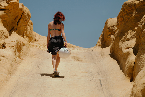 Young redhead woman with a motorcycle helmet wearing in black wrap and swimwear walking across arid path
