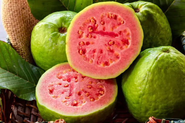 Closeup of a red guava cut in half, in the background several guavas and green leaf
