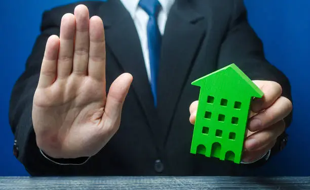 Photo of Man with a green house makes a stop gesture. Bank refusal to provide a mortgage loan. Housing problems, unscrupulous dishonest company builder. Confiscation pledged property. Refusal residence permit.