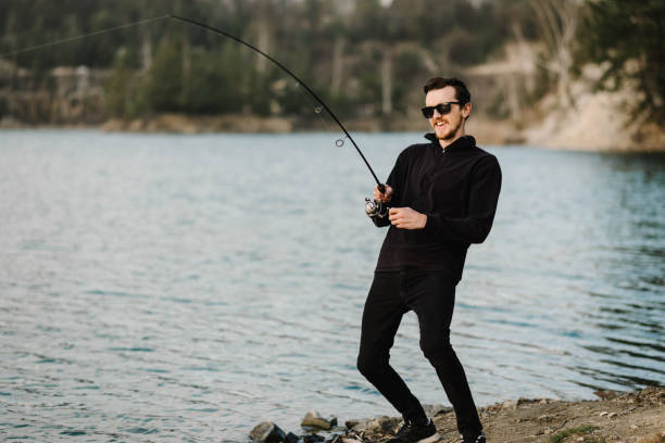 istock Fisherman with rod, spinning reel on the river bank. Man catching fish, pulling rod while fishing from lake or pond with text space at weekend. Fishing for pike, perch, carp on beach lake or pond. 1224641634