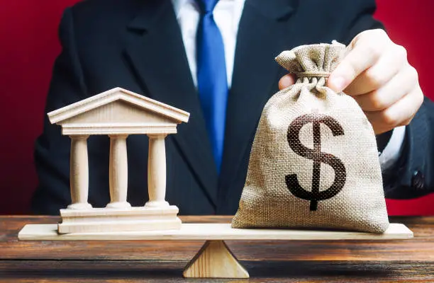 Photo of A man puts a money bag on scales opposite to building of government, bank, university. Budget and funding for normal functioning. Lobbying interests. Payment of taxes. Deposits and loans.