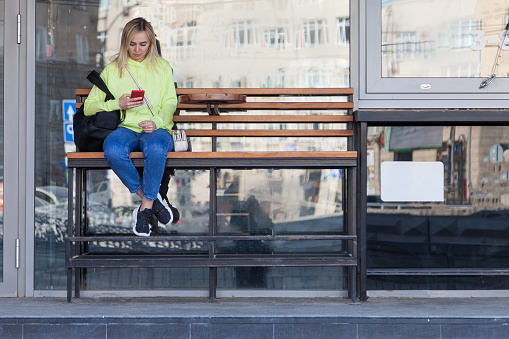 A blonde girl in a bright yellow sweatshirt sits on a wooden bench with a backpack near a coffee shop, holds a phone in her hands and waits for her order. The girl at the bus stop.