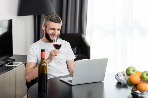 Selective focus of smiling freelancer holding glass of wine near laptop and fruits on table