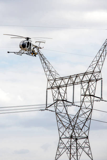 A repairman perches on a platform attached to a helicopter to work on a wire attached to a high voltage tower.