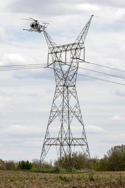 A repairman perches on a platform attached to a helicopter to work on a wire attached to a high voltage tower.