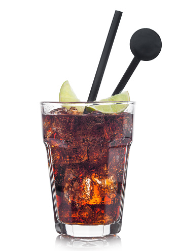Cuba Libre Cocktail in glass with ice cubes and slice of lime with black straw and stirrer on white background.