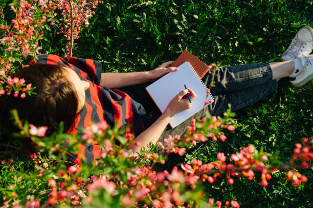 mädchen sitzt auf gras in einem blühenden garten mit einem notizblock und stift in den händen - nature writing women ideas stock-fotos und bilder