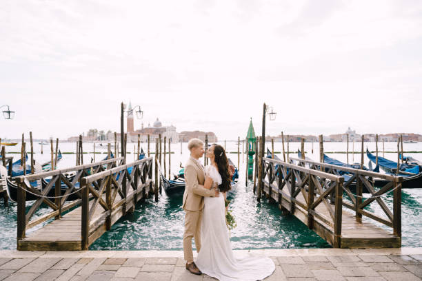 sposo e stand accanto al molo della gondola, abbracciandosi, a venezia, vicino a piazza san marco, con vista su san giorgio maggiore e sul cielo al tramonto. il più grande molo della gondola di venezia. - shoe groom wood luxury foto e immagini stock