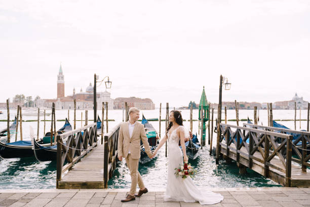 die braut und der bräutigam gehen entlang der gondelbrücke, halten hand in venedig, in der nähe der piazza san marco, mit blick auf san giorgio maggiore und den sonnenuntergang himmel. die größte gondelbrücke in venedig, italien. - shoe groom wood luxury stock-fotos und bilder