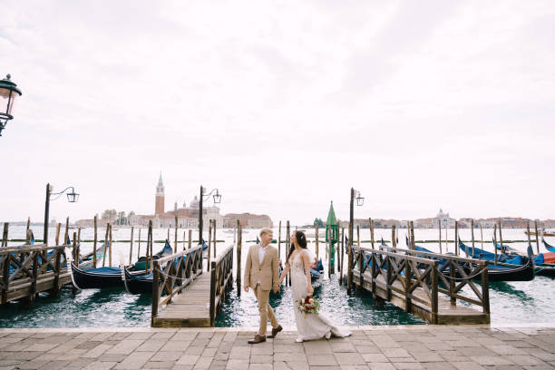 os noivos estão caminhando ao longo do píer de gôndola, segurando a mão em veneza, perto da piazza san marco, com vista para san giorgio maggiore e o céu do pôr do sol. o maior píer de gôndola em veneza, itália. - shoe groom wood luxury - fotografias e filmes do acervo