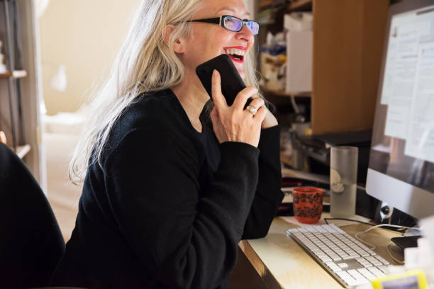Stylish 50+ woman working from home. Stylish 50+ woman working from untidy home office. She has long white hair and is wearing black casual clothes and red lipstick. She is using a computer and a mobile phone. Horizontal waist up indoors shot with copy space. quebec photos stock pictures, royalty-free photos & images