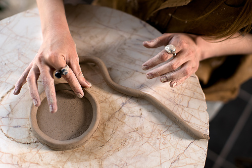 From above crop woman creating clay pot from coils on marble table in workshop