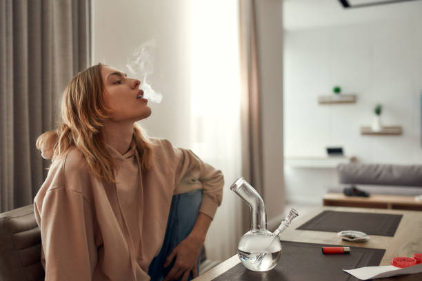 Relax, renew, revive. Young caucasian woman exhaling the smoke while smoking marijuana from a bong or glass water pipe, sitting in the kitchen. Red weed grinder and lighter on the table Young caucasian woman exhaling the smoke while smoking marijuana from a bong or glass water pipe, sitting in the kitchen. Red weed grinder and lighter on the table. Cannabis legalization concept bong stock pictures, royalty-free photos & images