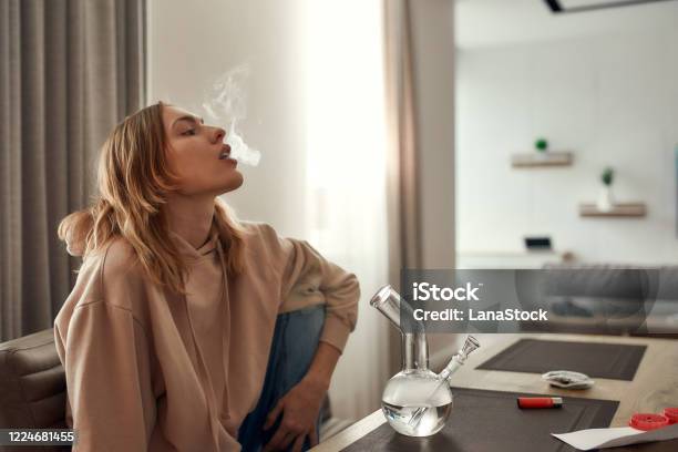 Relax Renew Revive Young Caucasian Woman Exhaling The Smoke While Smoking Marijuana From A Bong Or Glass Water Pipe Sitting In The Kitchen Red Weed Grinder And Lighter On The Table Stock Photo - Download Image Now