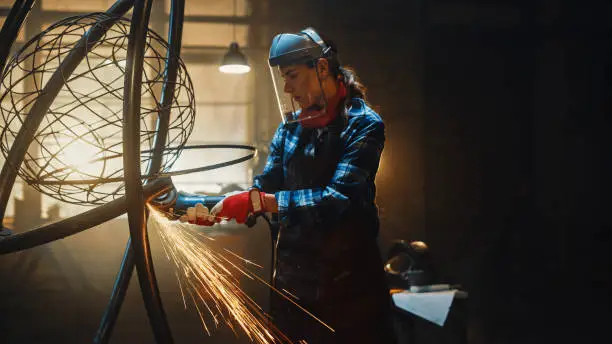 Photo of Beautiful Female Artist Uses an Angle Grinder to Make Brutal Metal Sculpture in Studio. Tomboy Girl Polishes Metal Tube with Sparks Flying Off It. Contemporary Fabricator Creating Abstract Steel Art.