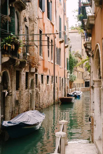 Photo of Boats moored at the walls of a building in a canal in Venice, Italy. Classic Venetian street views - wooden shutters, brick houses, bridges