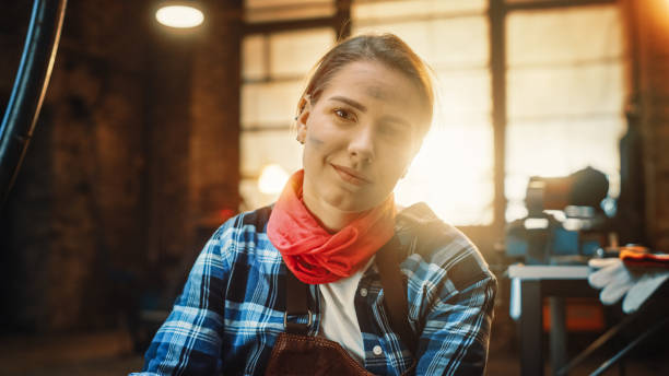 istock Young Beautiful Empowering Woman with Ear Piercing Gently Smiles at the Camera. Authentic Fabricator Wearing Work Clothes in a Metal Workshop. 1224682061