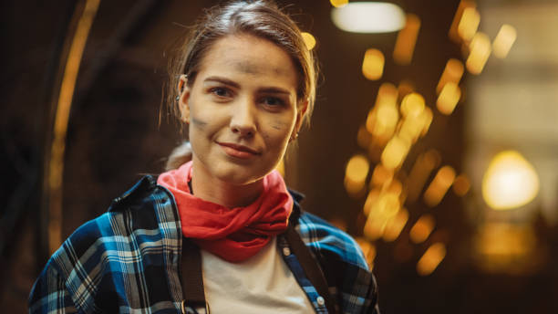 istock Young Beautiful Empowering Woman with Ear Piercing Gently Smiles at the Camera. Authentic Fabricator Wearing Work Clothes in a Metal Workshop. Sparks Flying on the Background. 1224682150