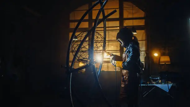 Photo of Talented Emerging Female Artist is Welding an Abstract, Brutal Metal Sculpture that Reflects the Present Moment. Beautiful Tomboy Fabricator in Checkered Shirt Creating Modern Steel Art.