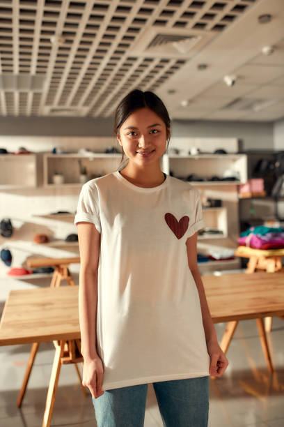 istock Design your presence. Young woman smiling at camera, while trying on printed t shirt, standing in the store. Clothes at custom T-shirt, clothing printing company 1224683062