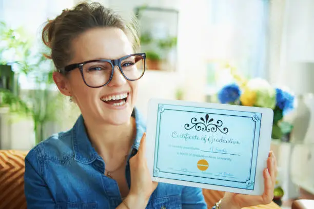 Portrait of smiling modern middle age housewife in jeans shirt in the modern living room in sunny day showing electronic Certificate of Graduation on tablet PC screen.