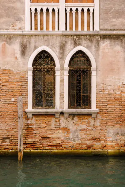 Photo of The facade of a corporatist apartment building in Venice, Italy. Buildings in waters of Venetian Canal. Typical Venetian windows with arches, columns and a sharp top, on the wall of a stone house.