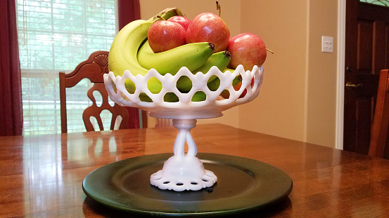 Fresh Ambrosia apples and green bananas in an antique milk glass fruit bowl in center of dining table.