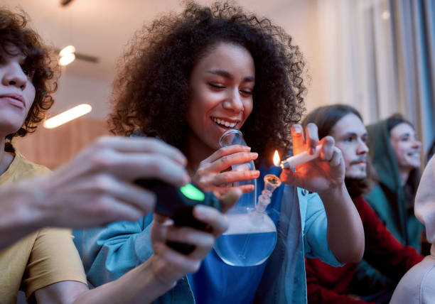 для отличного настроения. молодая афро-американская девушка зажигает марихуану в стеклянном бонге, отдыхая с друзьями на диване дома. моло� - video game friendship teenager togetherness стоковые фото и изображения