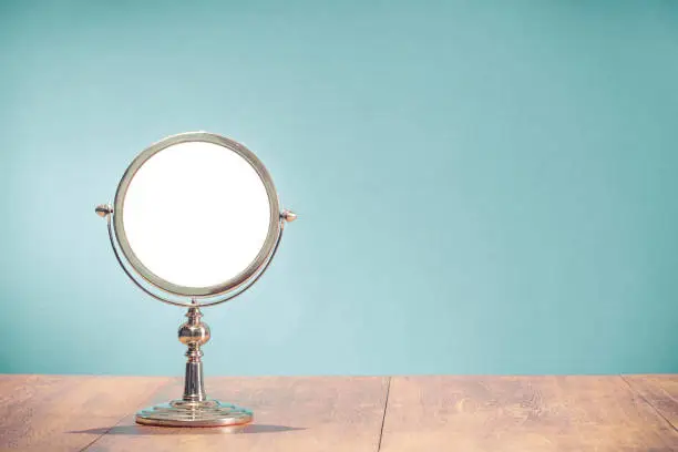 Photo of Retro classic silver colored round shape makeup mirror frame from circa 50s on aged oak wooden table front mint blue wall background. Nostalgic accessory. Vintage style filtered photo