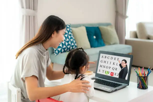Asian kindergarten school girl with mother video conference e-learning with teacher on laptop in living room at home. Homeschooling and distance learning ,online ,education and internet.
