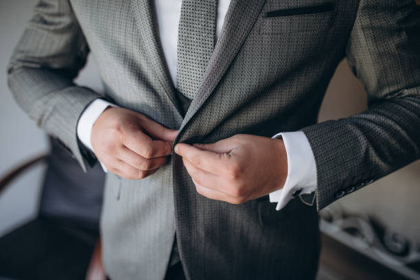 Young handsome stylish man dressed in modern formal clothes buttoning jacket. Close up of hands of guy in gray jacket, violet shirt. Person ready for wedding celebration or graduation.