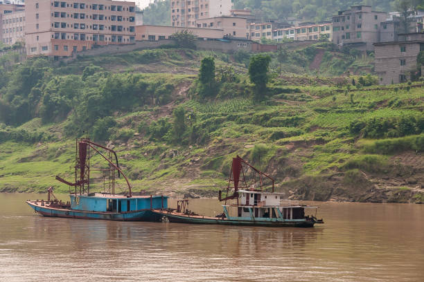 Fengdu, Chongqing, China - May 8, 2010: Yangtze River. Small rusty blue dredging boats on brown water neart green shoreline with agriculture plots and tall residential buildings.