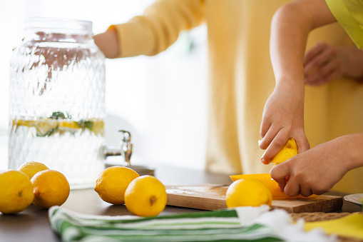 Cut out of young girl's arms, using lemon squeezer with a jar of lemonade and lemons placed on the kitchen table.