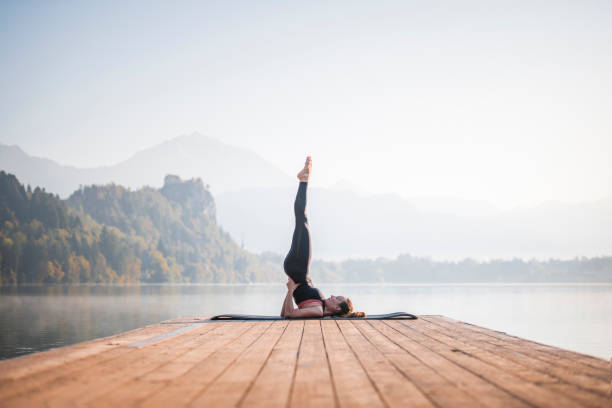 Woman in Supported Shoulder Stand Overlooking Lake Bled Side view of Caucasian woman in early 30s doing yoga Supported Shoulder Stand (Sarvangasana) on pier overlooking Lake Bled in morning. gorenjska stock pictures, royalty-free photos & images