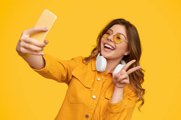 Cheerful lady gesturing V sign and taking selfie Happy stylish woman in sunglasses smiling and showing V sign while taking selfie against yellow background peace sign gesture photos stock pictures, royalty-free photos & images