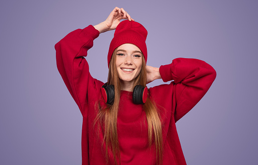Happy modern young woman with long ginger hair smiling and adjusting stylish red hat while standing against violet background