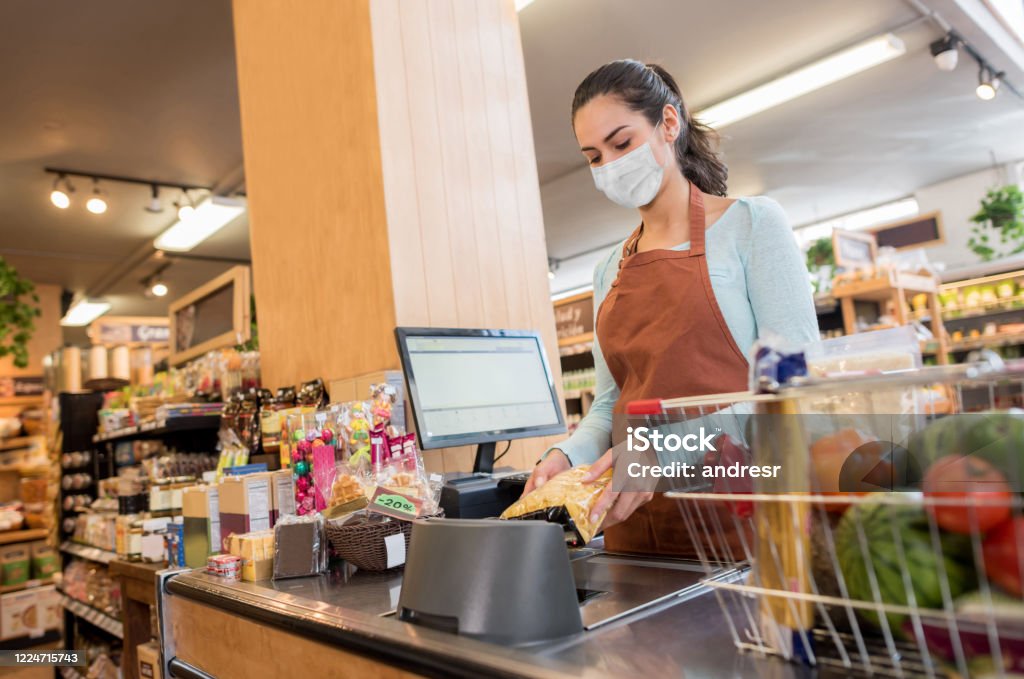 Cashier working at the supermarket wearing a facemask while scanning products Latin American cashier working at the supermarket wearing a facemask while scanning products â quarantine lifestyle concepts Supermarket Stock Photo