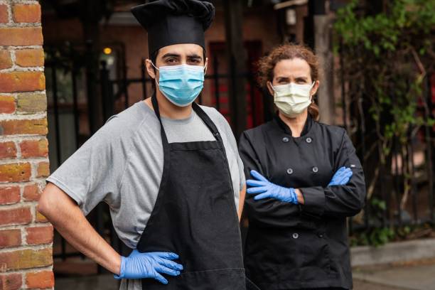 istock Young chef posing with female coworker wearing both a protective masks and gloves during a break looking at the camera 1224722879