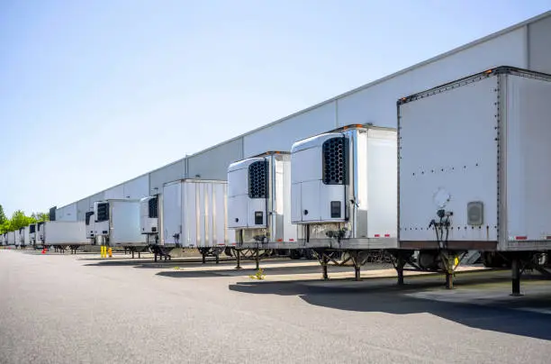 Industrial grade refrigerator and dry van semi trailers with reefer units on the front wall and without it standing at warehouse dock gates loading commercial cargo for next freight delivery