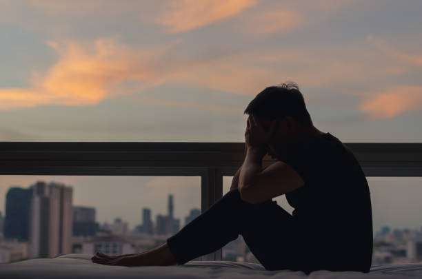 A person sitting and feeling depressed on bed. A person sitting and feeling depressed on bed with city view in dusk moment. Stay home, depression and loneliness concept. suicide photos stock pictures, royalty-free photos & images