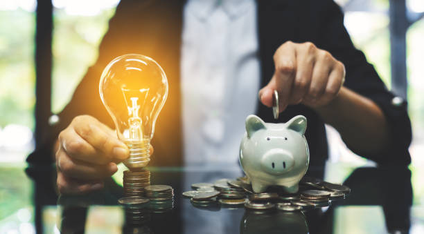 A businesswoman putting coin into piggy bank and a light bulb over coins stack on the table stock photo