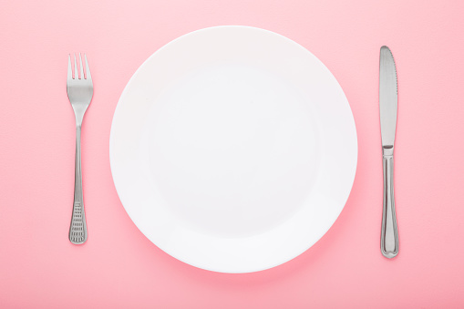 White plate with fork and knife on light pink table background. Pastel color. Closeup. Meal waiting concept. Top down view.