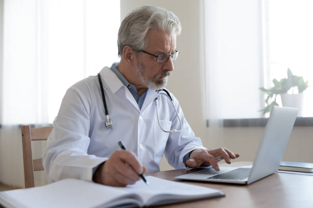 Serious senior physician using computer app making notes in journal Serious senior mature physician using computer app making notes in medical journal. Older adult doctor therapist consulting remote patient online by video call, watching healthcare webinar training. medical journal stock pictures, royalty-free photos & images